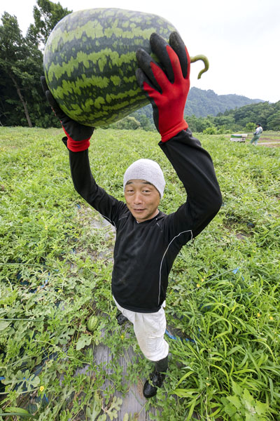 「寿風土ファーム」の小林栄一さんと「戦水甘（せんすいかん）」シリーズのラグビーボールのようなスイカ（提供写真）